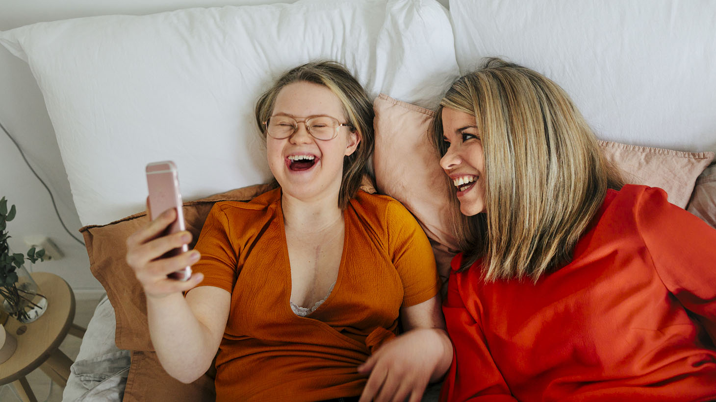 Två kvinnor i orange tröjor tittar på något i en mobil och skrattar.